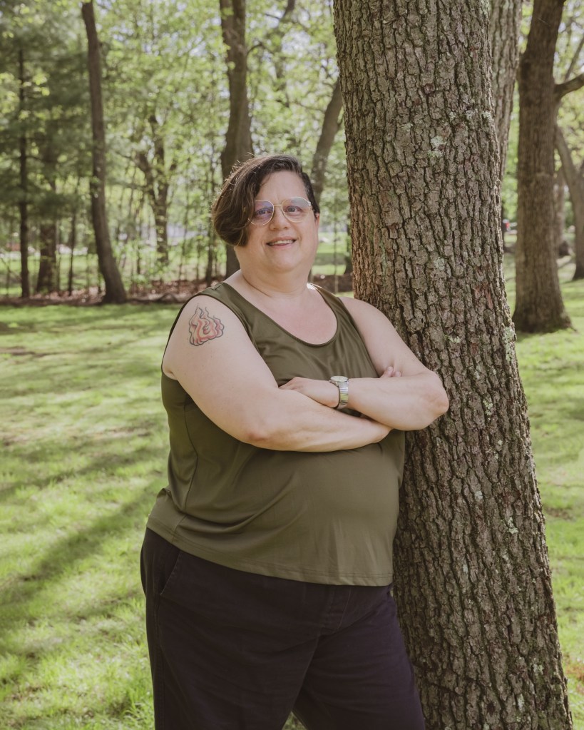 Headshot of Rachel. They are leaning against a tree with their arms folded, looking into the camera.